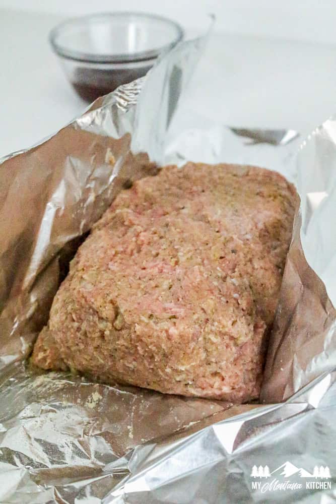 Side view of raw turkey meatloaf loaf shaped in foil, with BBQ sauce in the background