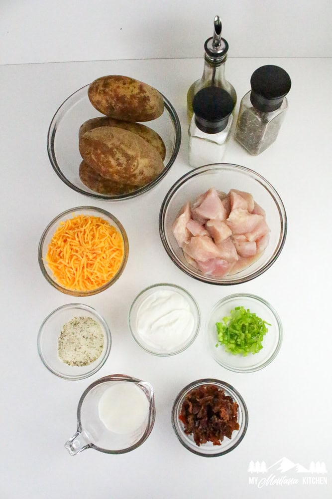 Flat lay of all casserole ingredients including raw potatoes, shredded cheese, chopped bacon, chicken, ranch seasoning, sour cream, and green onions