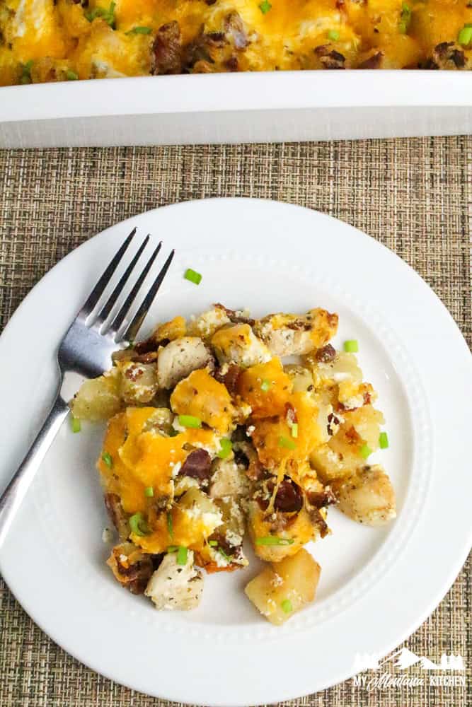 Photo of a serving of cheesy chicken, bacon, and potatoes on a white plate with a fork and green onions sprinkled on top