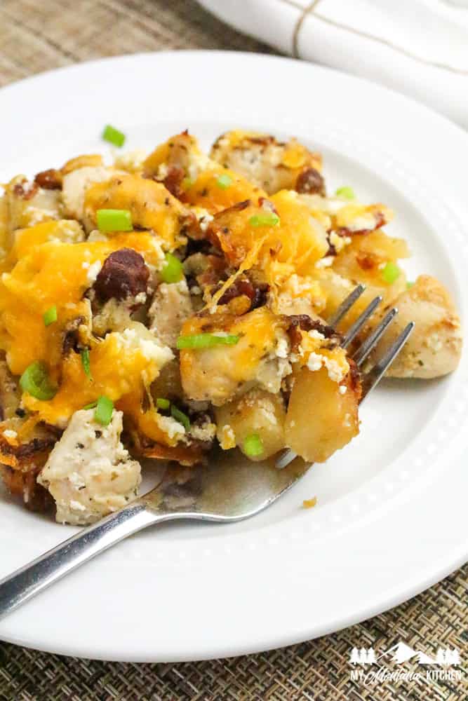 Photo of a serving of cheesy chicken, bacon, and potatoes on a white plate with a fork and green onions sprinkled on top