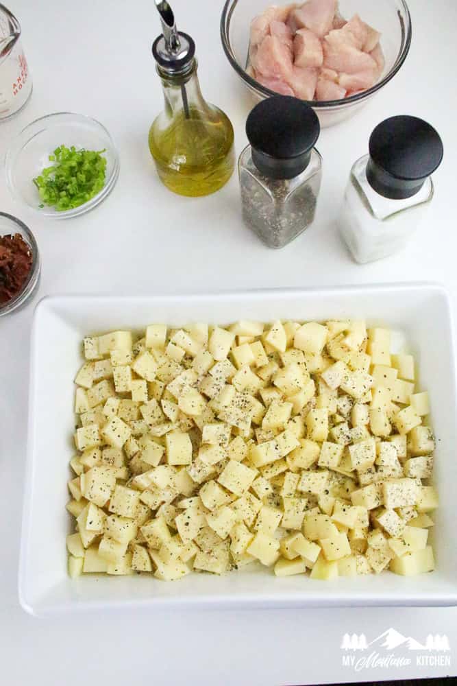 Flat lay of cubed raw potatoes in a casserole dish, surrounded by bowls of chopped chicken, bacon bits, oil, seasoning, and green onions