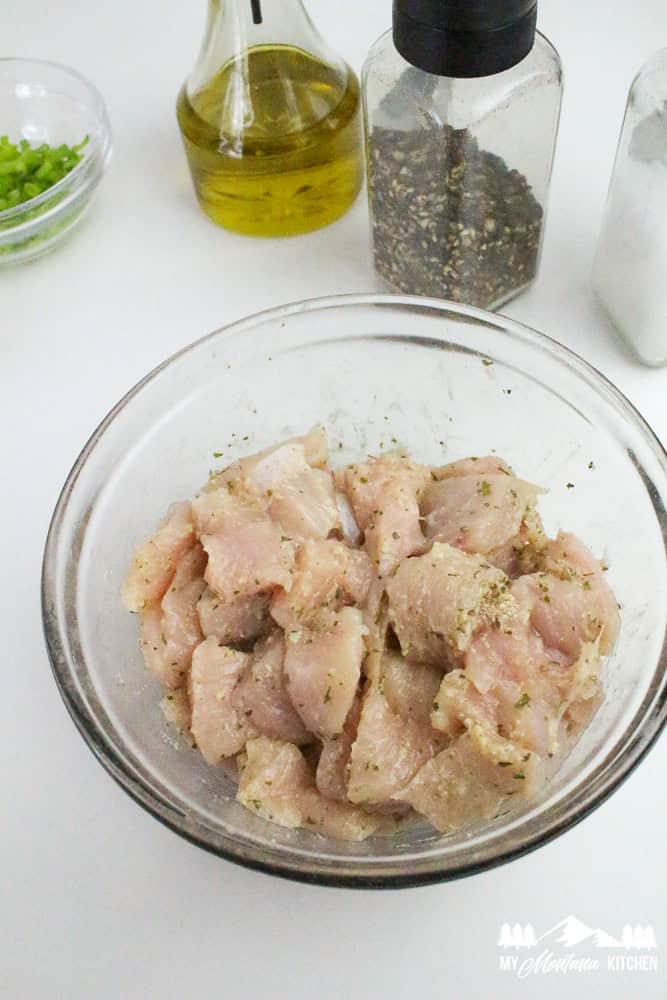 Bowl of raw, seasoned boneless skinless chicken surrounded by oil, pepper, salt, and chopped green onions on a white background