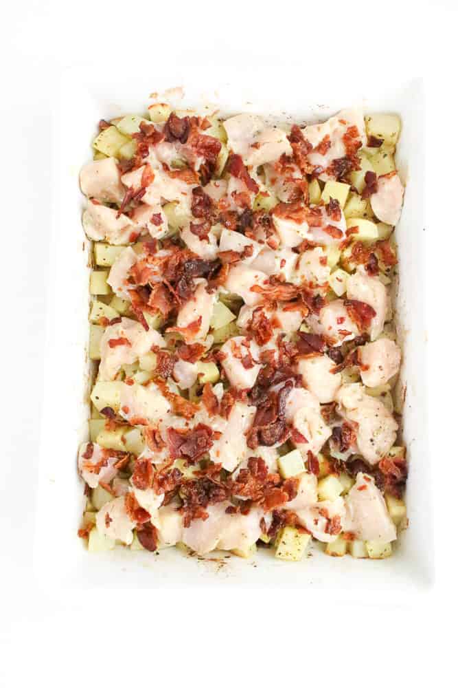 Mid-prep image of diced chicken and bacon layered over seasoned potatoes in a white casserole dish