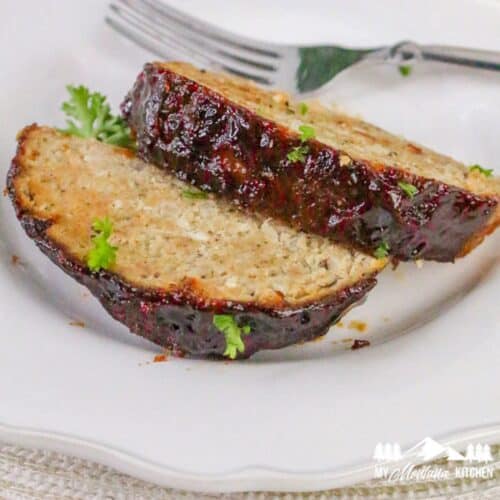 Close-up of two thick slices of air fryer turkey meatloaf topped with ketchup glaze, garnished with fresh parsley on a white plate.