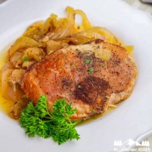 Close-up of a fully cooked seasoned pork chop served with tender apples and onions on a white plate, garnished with parsley