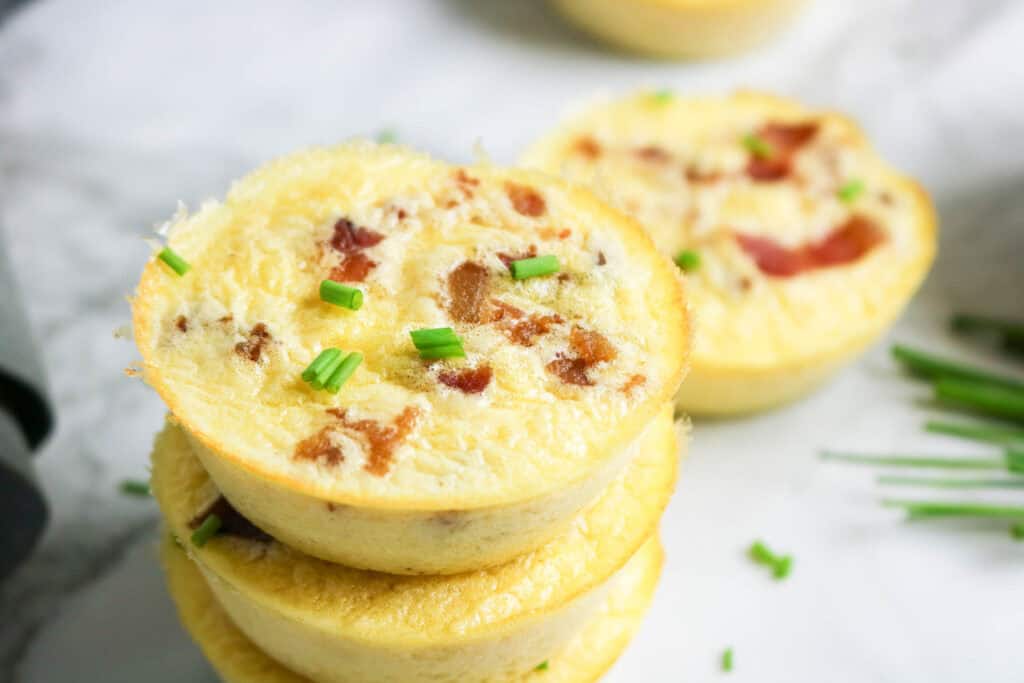 Stacked cottage cheese egg bites topped with crispy bacon and fresh chives on a marble surface.