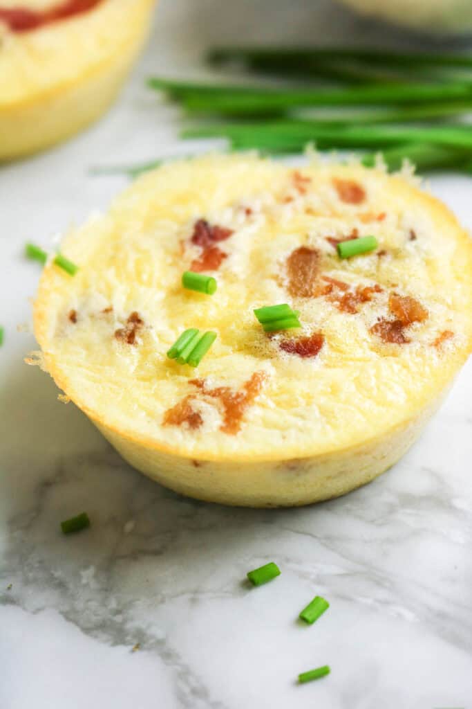 Close-up of baked cottage cheese egg bite topped with bacon and chives, bowl of bacon in background.
